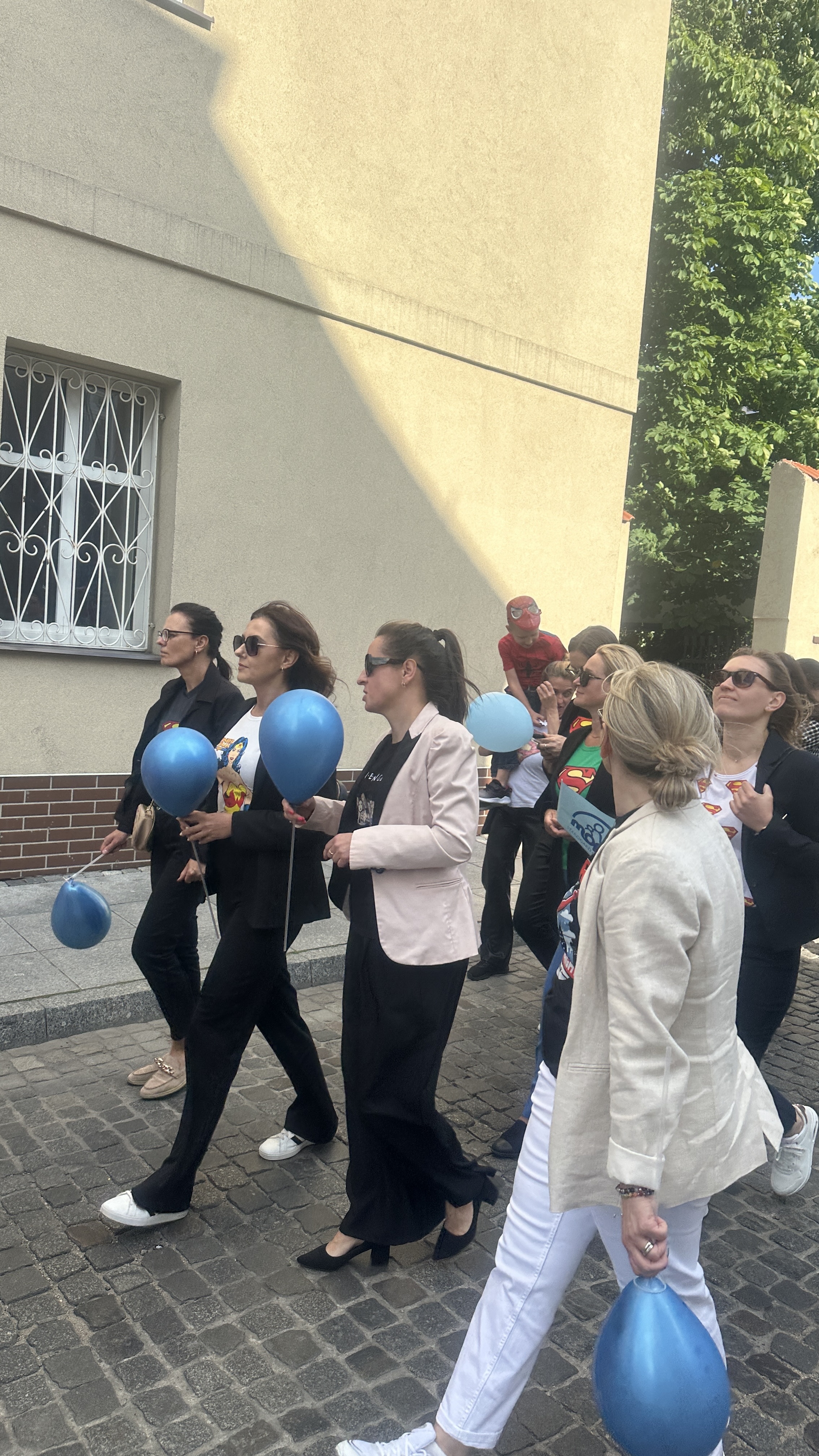 Grupa osób idąca kostką brukową (ulicą) trzyma w ręce niebieskie balony.